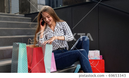 Girl sitting on stairs with bags talking on smartphone about sale in shopping mall in Black Friday Girl sitting on stairs with bags talking on smartphone about sale in shopping mall in Black Friday 69653343