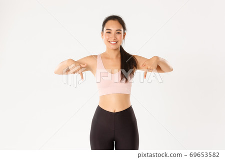 Sport, wellbeing and active lifestyle concept. Smiling fit and slim female fitness instructor, asian girl workout in gym and showing something, pointing fingers down, standing white background Sport, wellbeing and active lifestyle concept. Smiling fit and slim female fitness instructor, asian girl workout in gym and showing something, pointing fingers down, standing white background 69653582