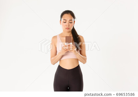 Sport, wellbeing and active lifestyle concept. Indecisive cute asian girl in workout clothing, looking hesitant at chocolate bar, trying resist temptation as losing weight on diet 69653586
