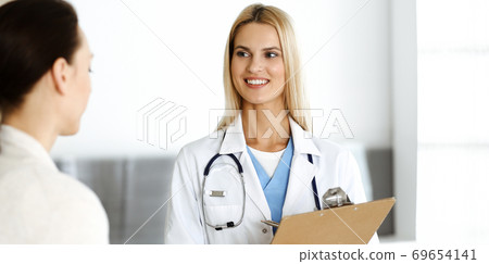 Woman-doctor at work in hospital is happy to consult female patient. Blonde physician checks medical history record and exam results while using clipboard. Medicine concept 69654141
