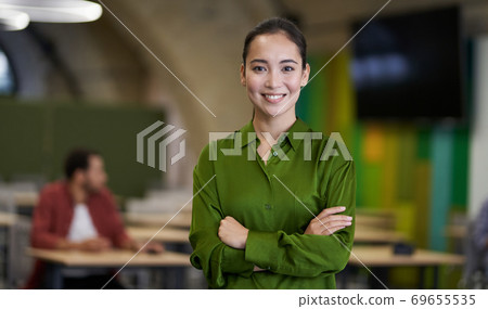Portrait of a young beautiful female office worker keeping arms crossed, looking at camera and smiling while standing in the modern coworking space Portrait of a young beautiful female office worker keeping arms crossed, looking at camera and smiling while standing in the modern coworking space 69655535