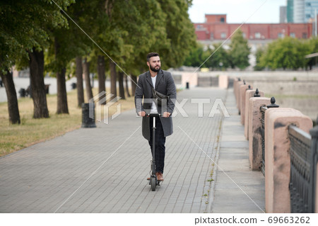 Contemporary young bearded businessman in coat riding on electric scooter Contemporary young bearded businessman in coat riding on electric scooter 69663262