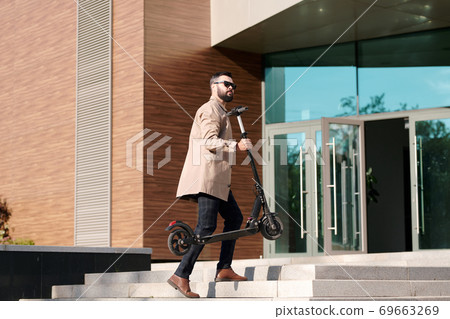 Young businessman in trenchcoat, pants and sunglasses carrying electric scooter 69663269
