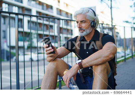 Portrait of mature man with headphones sitting outdoors in city, using smartphone. Portrait of mature man with headphones sitting outdoors in city, using smartphone. 69664441