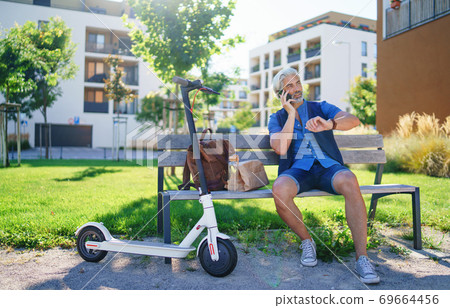 Portrait of mature man with electric scooter sitting outdoors in city, using smartphone. Portrait of mature man with electric scooter sitting outdoors in city, using smartphone. 69664456