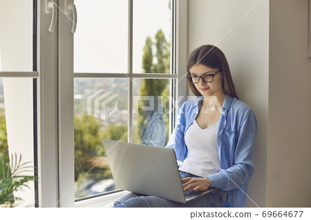 Working from home. Young girl using laptop for online video call or web conference with boss while sitting on windowsill Working from home. Young girl using laptop for online video call or web conference with boss while sitting on windowsill 69664677
