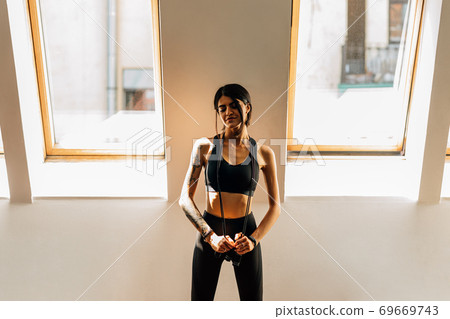 Slim sportswoman standing between two windows in gym with skipping rope and resting Slim sportswoman standing between two windows in gym with skipping rope and resting 69669743