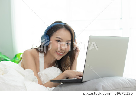 Asian woman working on a laptop sitting on the bed in the house. Asian woman working on a laptop sitting on the bed in the house. 69672150