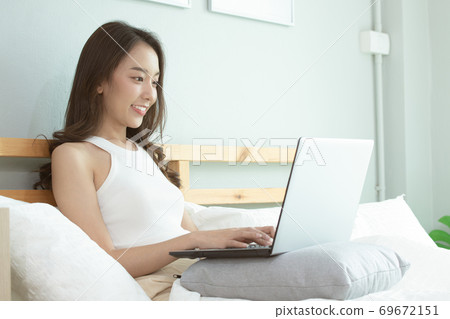 Asian woman working on a laptop sitting on the bed in the house. Asian woman working on a laptop sitting on the bed in the house. 69672151
