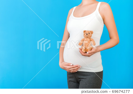 Close up of pregnant woman in home clothing holding teddy bear against her abdomen at blue background. Parenthood concept. Copy space Close up of pregnant woman in home clothing holding teddy bear against her abdomen at blue background. Parenthood concept. Copy space 69674277