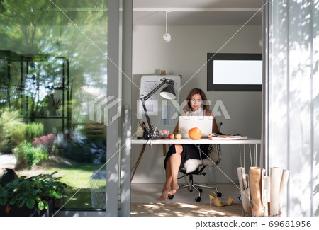 Mature woman working indoors in home office in container house in backyard. Mature woman working indoors in home office in container house in backyard. 69681956