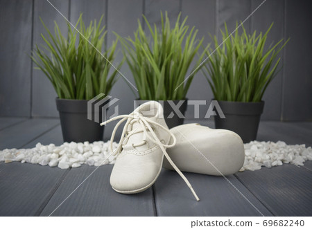 Newborn baby boy's booties in white, with decorative stones, green plants and a gray wooden background. Children's shoes 69682240