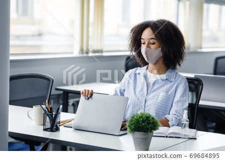 End of day and go home. African american woman in protective mask closes laptop, sitting at table with antiseptic in modern office interior End of day and go home. African american woman in protective mask closes laptop, sitting at table with antiseptic in modern office interior 69684895