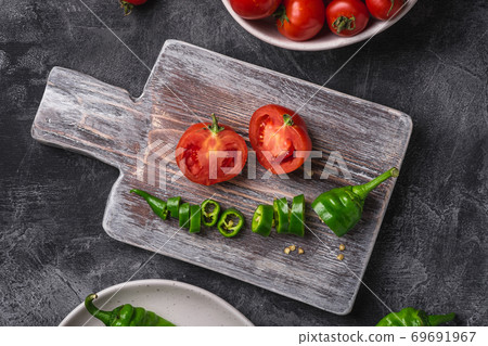 Fresh ripe tomatoes in bowl near to old wooden cutting board with hot chili peppers in plate, dark stone concrete background, top view close up Fresh ripe tomatoes in bowl near to old wooden cutting board with hot chili peppers in plate, dark stone concrete background, top view close up 69691967