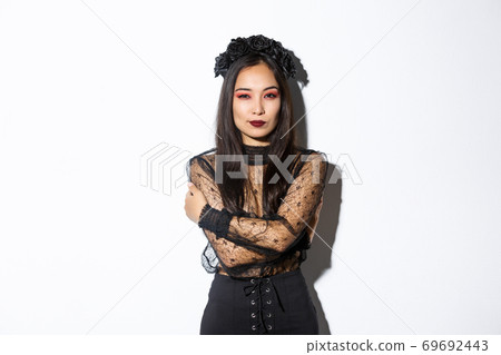 Young elegant woman in stylish gothic dress and wreath, embracing her body and looking sassy at camera, wearing halloween costume of undead widow, standing over white background 69692443