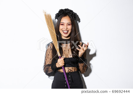 Image of witch in gothic lace dress and black wreath making evil laugh and holding broom, celebrating halloween, standing over white background Image of witch in gothic lace dress and black wreath making evil laugh and holding broom, celebrating halloween, standing over white background 69692444
