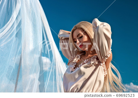 Portrait of a beautiful girl, blonde with long hair in a field, on a background of white transparent fabric 69692523