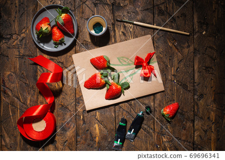 Gift card with live strawberries. On the table are satin ribbon, paintbrush and tubes of paint. Top Gift card with live strawberries. On the table are satin ribbon, paintbrush and tubes of paint. Top 69696341