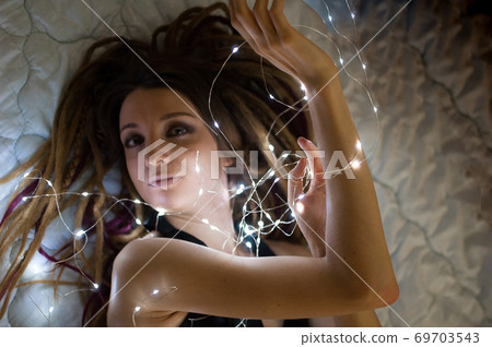 Indoors female portrait of a woman with dreadlocks hairstyle and smoky eyes makeup holding lights garland near her face, femininity and hollidays concepts Indoors female portrait of a woman with dreadlocks hairstyle and smoky eyes makeup holding lights garland near her face, femininity and hollidays concepts 69703543