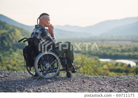 Young pensive man in a wheelchair enjoying the beauty of nature in the mountains. Rehabilitation after injury. Young pensive man in a wheelchair enjoying the beauty of nature in the mountains. Rehabilitation after injury. 69711175