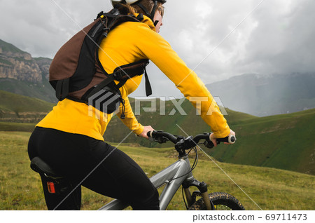 A young woman in a helmet and with a backpack stands with a bicycle and looks at the mountains in cloudy weather. Mountain mtb bike 69711473