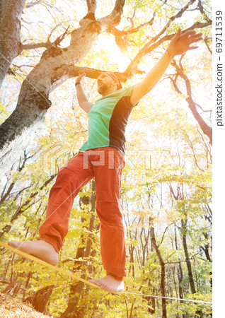 A bearded man in age walks along a slackline stretched in an autumn forest. Active recreation for middle-aged people A bearded man in age walks along a slackline stretched in an autumn forest. Active recreation for middle-aged people 69711539