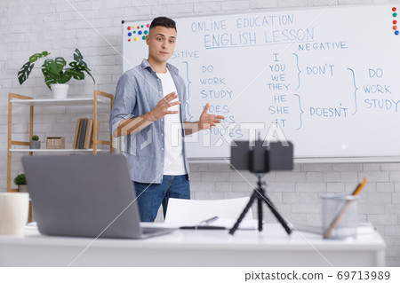 Remote education at home during epidemic. Young man explains rules of English for students, standing near blackboard and looks in webcam of smartphone on table 69713989