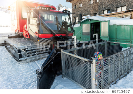 Cute adorable playful happy toddler boy inside red modern snowcat ratrack snowplow box grooming standing on peak alpine skiing resort Ischgl Austria. Heavy machinery mountain equipment track vehicle 69715497
