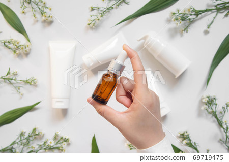 Serum or essential oil w/ organic herbal extract. Woman's hand holding brown glass vial w/ dropper and natural skincare bottles on white background. Natural beauty cosmetic product. Top view. Serum or essential oil w/ organic herbal extract. Woman's hand holding brown glass vial w/ dropper and natural skincare bottles on white background. Natural beauty cosmetic product. Top view. 69719475