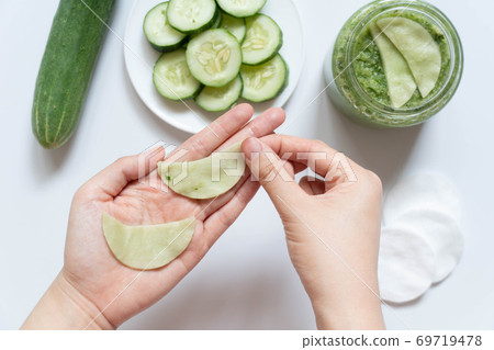 Cucumber eyes mask or makeup remover pads made w/ natural ingredients for healthy skin care. Hand holding DIY eyes mask w/ cucumber, facial cotton pads and jar on white table. Homemade beauty product. Cucumber eyes mask or makeup remover pads made w/ natural ingredients for healthy skin care. Hand holding DIY eyes mask w/ cucumber, facial cotton pads and jar on white table. Homemade beauty product. 69719478