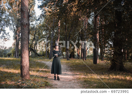 Pretty young beautiful woman in dark dress and witch hat standing in middle of autumn woods or park. Halloween party costume. Portrait in full growth. Shadow on the face. Pretty young beautiful woman in dark dress and witch hat standing in middle of autumn woods or park. Halloween party costume. Portrait in full growth. Shadow on the face. 69723991