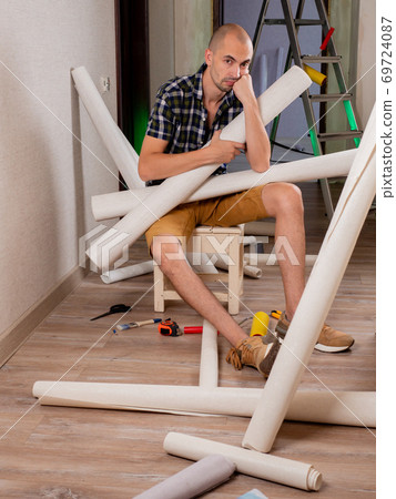 Renovation of the house. Preparation for wallpapering. A sad man sits on a bench and holds rolls of Wallpaper in his hands. Renovation of the house. Preparation for wallpapering. A sad man sits on a bench and holds rolls of Wallpaper in his hands. 69724087