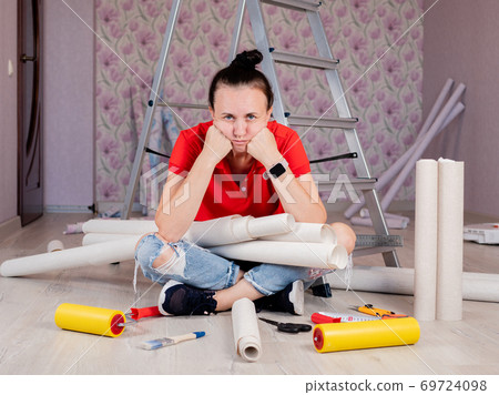 Preparing for wallpapering in the room. A sad woman is sitting on the floor with rolls of Wallpaper in her hands. Preparing for wallpapering in the room. A sad woman is sitting on the floor with rolls of Wallpaper in her hands. 69724098