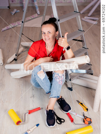 Portrait of a woman sitting on the floor with rolls of Wallpaper in her hands on the background of a stepladder. Portrait of a woman sitting on the floor with rolls of Wallpaper in her hands on the background of a stepladder. 69724102
