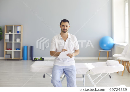 Young male doctor with clipboard sitting on hospital bed in office of new clinic Young male doctor with clipboard sitting on hospital bed in office of new clinic 69724889