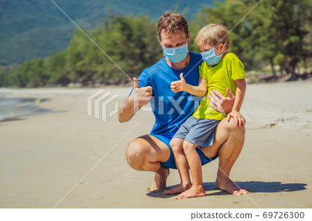 Father squatting holding son playing fists with thumbs up, wearing face blue mask protect coronavirus walking nature sand sea beach near forest park sun day. COVID Global pandemic virus reality 69726300