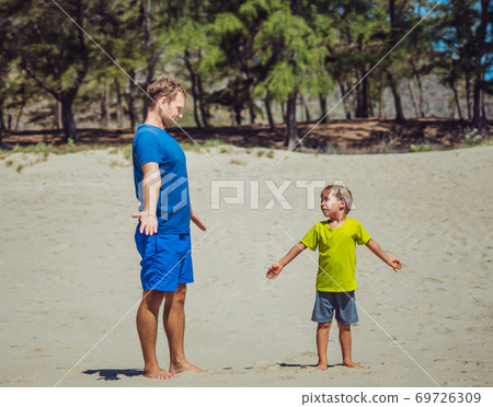 Sport for little children. Father cute son exercising on sand beach near forest park, having workout enjoying together in the fresh air, hand exercises technique, daddy influence on boy worldview Sport for little children. Father cute son exercising on sand beach near forest park, having workout enjoying together in the fresh air, hand exercises technique, daddy influence on boy worldview 69726309