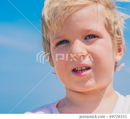 Close-up boy portrait . Serious male child squints a little in sunny weather looks into the distance. Clear blue sky background. 5 years old, freckles face, blue eyes, light t-shirt. Children concept 69726331