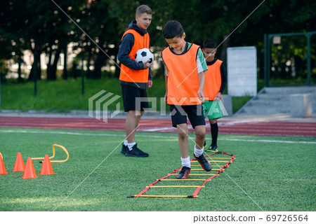 Two school boys are running ladder drills on the turf during football summer camp. Intense soccer training with coach. 69726564