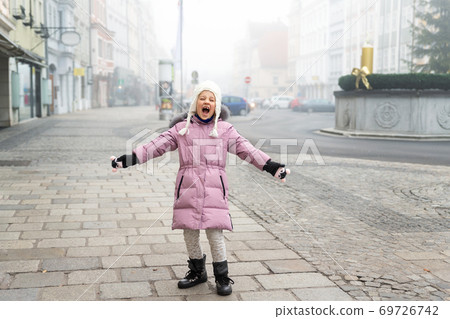 Cute liitle sad lost caucasian girl in warm long pink jacket and hat shouting loud on empty street of old european city Wels. Screaming angry alone scared child in town in Morning fog on backround 69726742