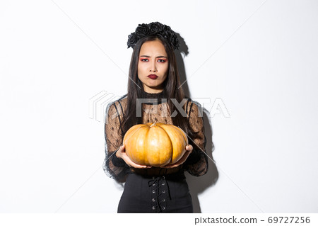 Image of grimacing asian woman in witch costume hate carve pumpkin for halloween, looking disappointed, standing over white background Image of grimacing asian woman in witch costume hate carve pumpkin for halloween, looking disappointed, standing over white background 69727256