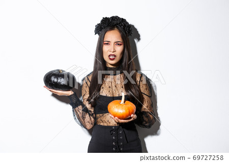 Portrait of disappointed frowning girl in witch costume, holding pumpkins and complaining, grimacing while standing over white background Portrait of disappointed frowning girl in witch costume, holding pumpkins and complaining, grimacing while standing over white background 69727258