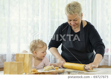 Beautyful smilling little girl helps her grandmother roll out the cookie dough. Beautyful smilling little girl helps her grandmother roll out the cookie dough. 69727873