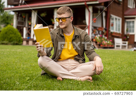 Hipster guy resting on lawn with paper book and cup of tea, reading outdoors Hipster guy resting on lawn with paper book and cup of tea, reading outdoors 69727903