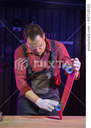 middle-aged man in jumpsuit testing plastic kids skateboard in carpentry middle-aged man in jumpsuit testing plastic kids skateboard in carpentry 69728010
