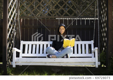 Caucasian girl in hoodie sits with book in garden on swing and broadly smiles. 69728073