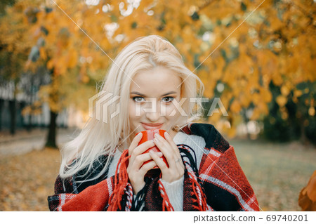 Beautiful blonde Woman with Autumn Leaves on Fall Nature Background. Alone brooding woman in white sweater and red plaid holding cup and enjoys nature 69740201