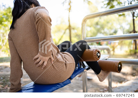 Sad asian woman with back ache while practicing fitness,injury to the back muscles, female people is suffering from a back pain and touch her back after exercising in a sit up position in oudtoor park 69747245