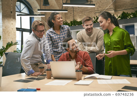 Group of young happy multicultural business people looking at laptop screen and discussing work or project results while standing in the modern office or coworking space 69748236