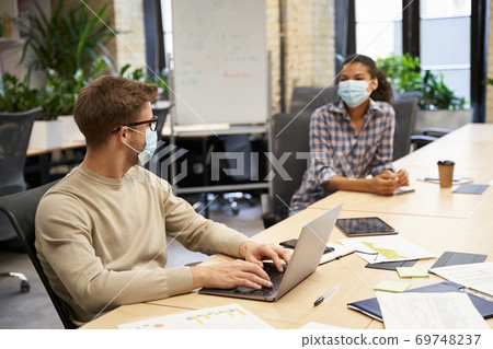 Two young coworkers wearing medical protective masks sitting together at the table in the modern office and talking, guy working on laptop Two young coworkers wearing medical protective masks sitting together at the table in the modern office and talking, guy working on laptop 69748237